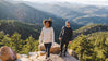 Two people wearing lightweight alpaca crew sweaters walking on a mountain trail with a valley and forested hills in the background