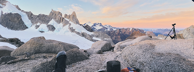 Hiker’s boot and camping gear on granite boulders, tripod set up, sunrise over snow‑capped jagged peaks