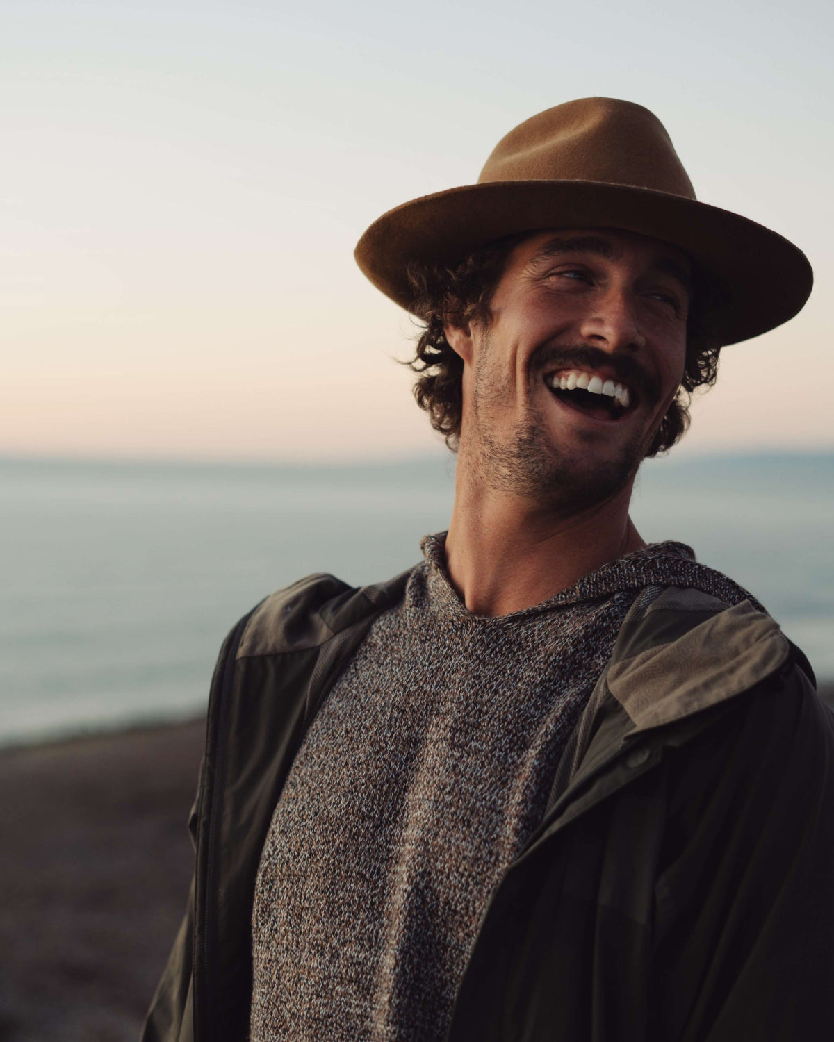 A smiling man wearing a hat and The Vida hoodie in the beach
