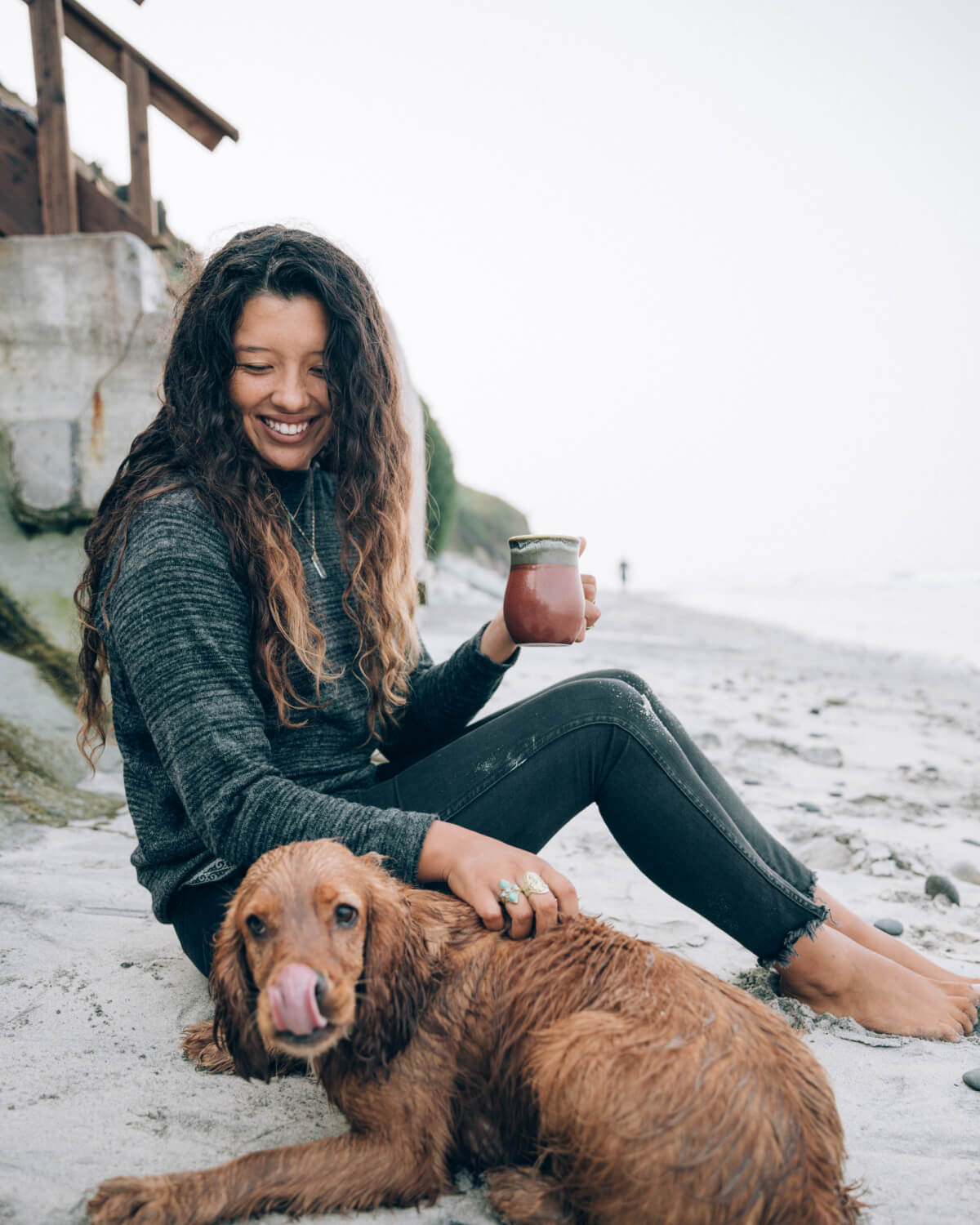 Woman Named Maya in The Hoodie Charcoal / Dark Grey Hooded Sweater Petting Her Dog and Holding a Mug on a Beach – PAKA Apparel