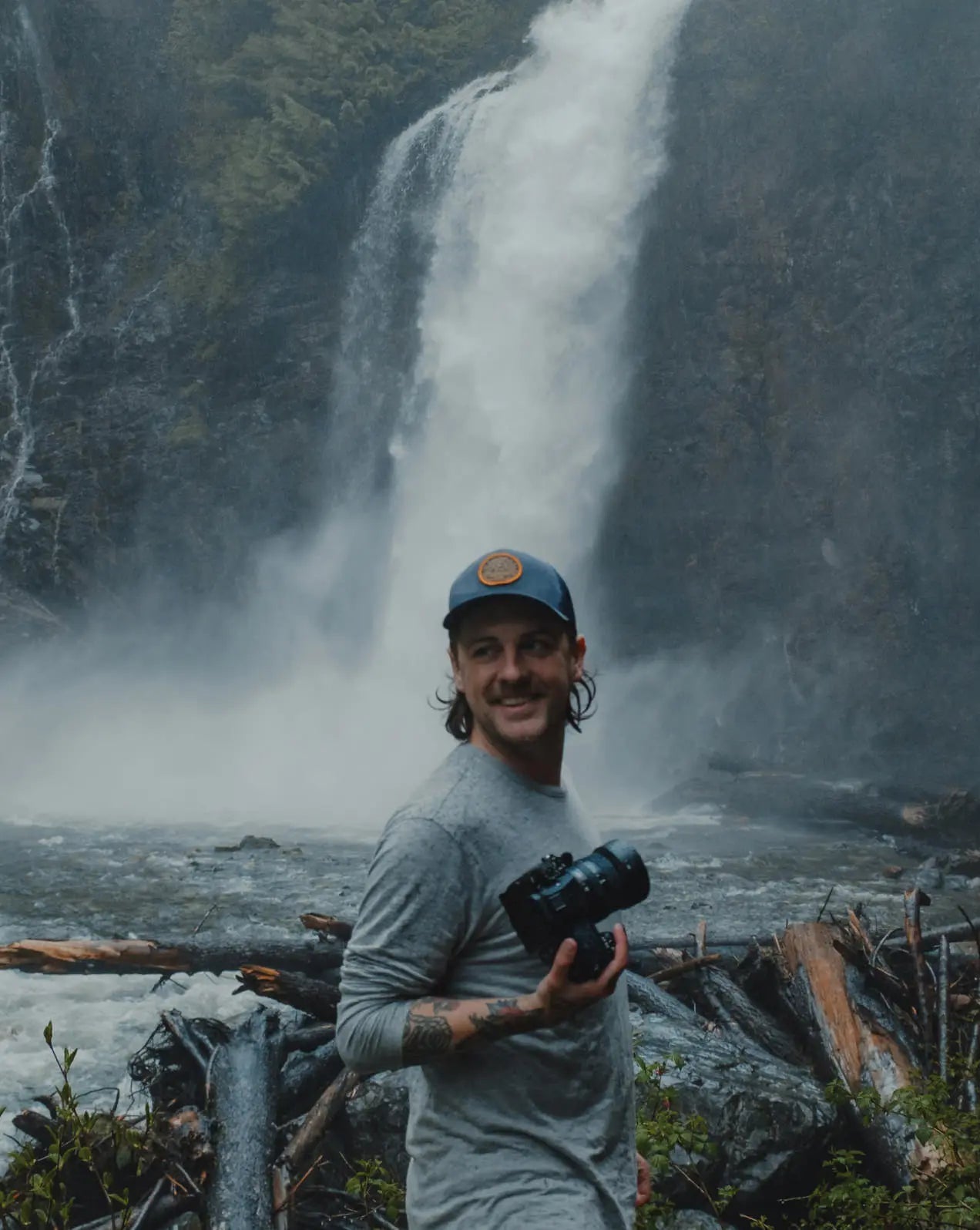 Man Named Mio Smiling with a Camera in Front a Waterfall Wearing Heather Grey Aire Long Sleeve Shirt – PAKA Apparel