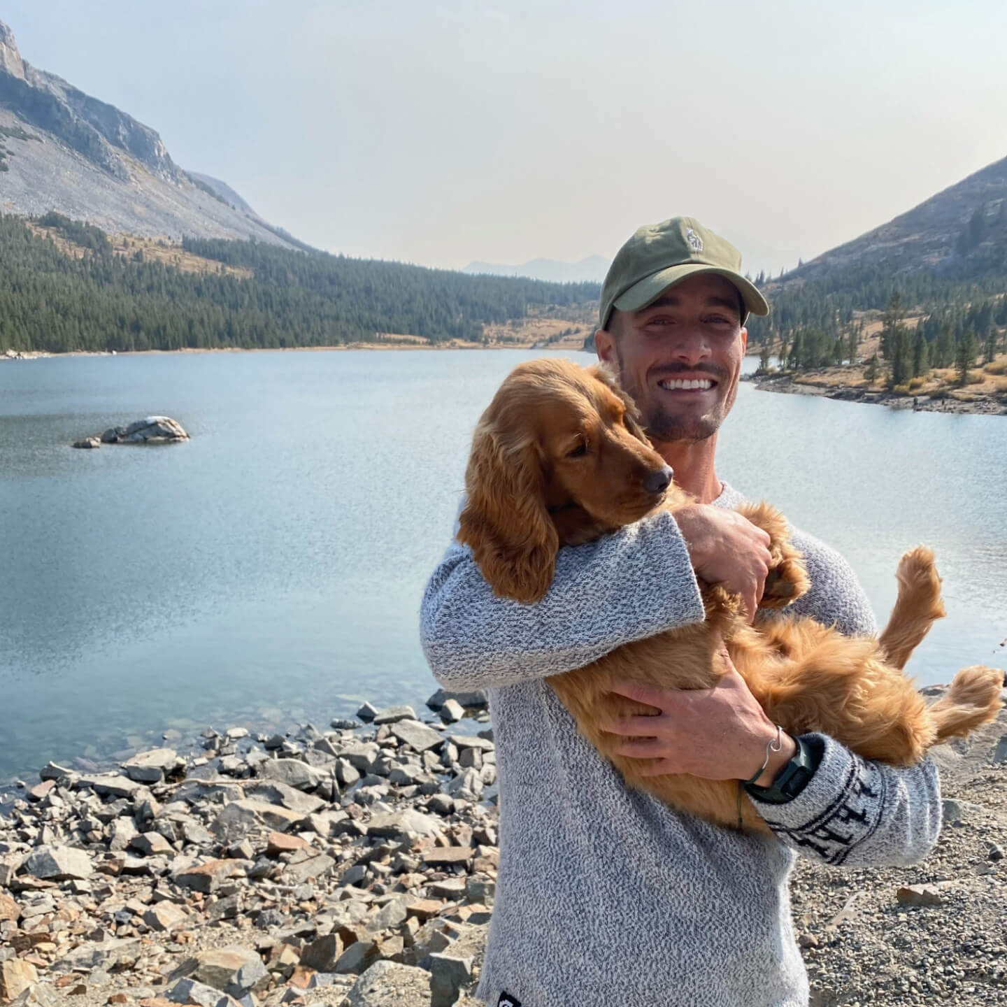 Man wearing a light gray unisex Costa alpaca crewneck sweater holding a brown dog on a rocky mountain lake shore