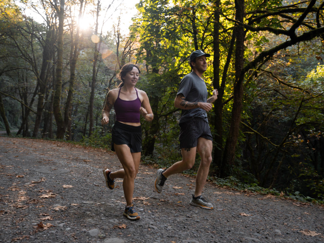 Two runners jogging on a forest trail wearing ankle socks and running shoes in dappled sunlight