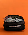 Front view of black upcycled packing cube open with clothes inside, showing mesh top pocket and woven handle on orange background