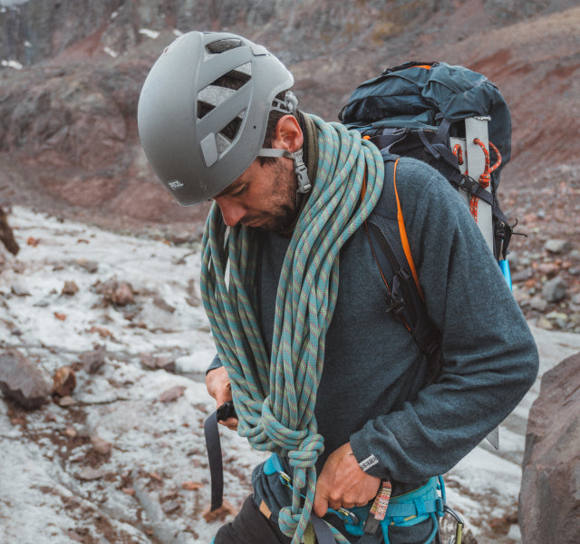 Climber adjusting a harness while wearing a dark gray ultralight crew sweater, helmet, rope, and backpack on a rocky glacier