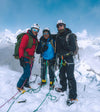 Three climbers standing on a snowy mountain summit with ropes and ice axes, snow-capped peaks and blue sky behind them