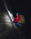 Climber wearing a red jacket and green backpack ascending a steep rock face at night with a headlamp and rope
