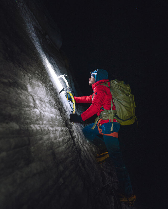 Climber wearing a red jacket and green backpack ascending a steep rock face at night with a headlamp and rope