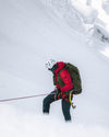 Climber rappelling down a steep snowy slope wearing a red puffer jacket and green backpack with rope and snowy mountain backdrop