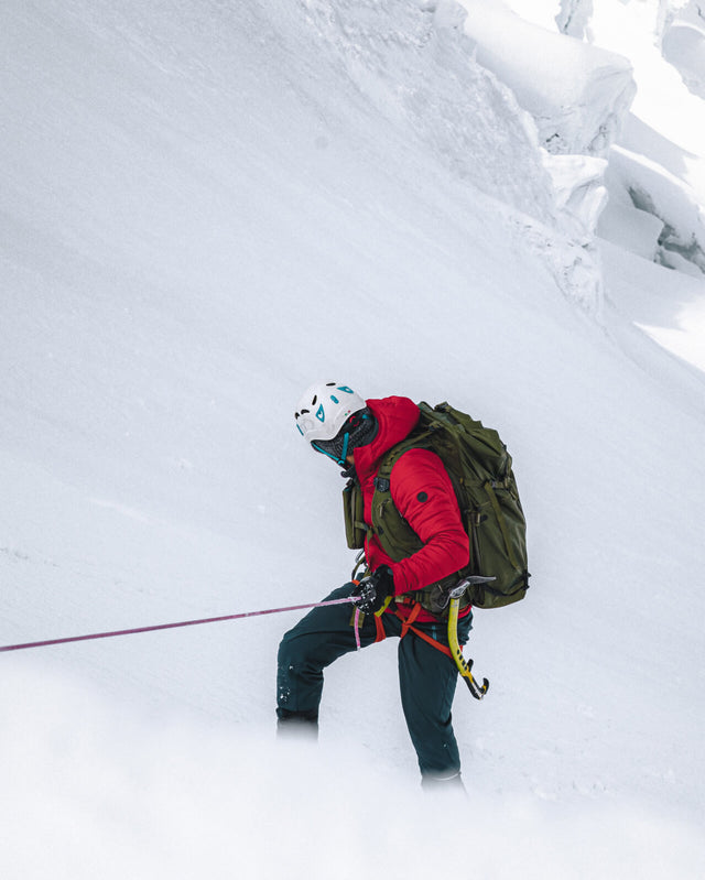Climber rappelling down a steep snowy slope wearing a red puffer jacket and green backpack with rope and snowy mountain backdrop