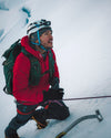 Mountaineer kneeling on a snowy slope during an ascent, wearing a red jacket and helmet, holding an ice axe with glacier and cloudy sky behind
