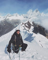 Climber kneeling on a snowy ridge holding ice axes with jagged Cordillera Blanca peaks behind