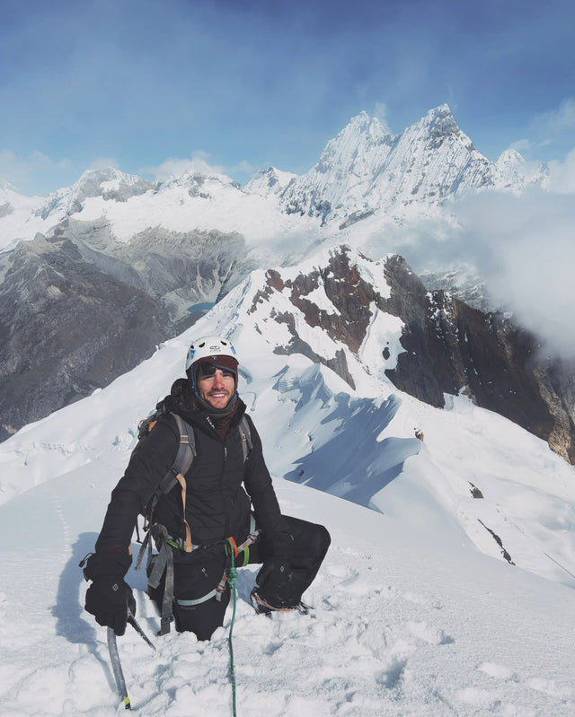 Climber kneeling on a snowy ridge holding ice axes with jagged Cordillera Blanca peaks behind