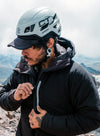 Man zipping a black insulated puffer jacket while wearing a climbing helmet on a rocky mountain ridge