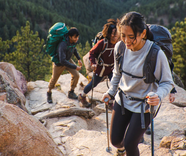 Woman leading two hikers up a rocky mountain trail using trekking poles, backpacks and forested peaks in the background