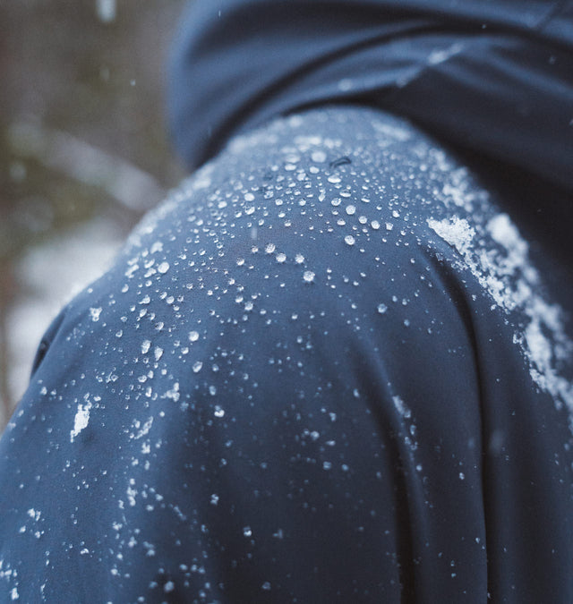 Close-up rear shoulder of navy APU jacket showcasing PAKAFILL 2.0 water beading rainproof shell with snow on the fabric