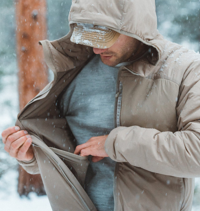 Front-quarter view of PAKAFILL insulated hooded jacket showcasing interior zip pocket with snowy forest background