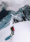 Mountaineer in a red jacket climbing a snowy mountain ridge toward jagged peaks under a cloudy sky