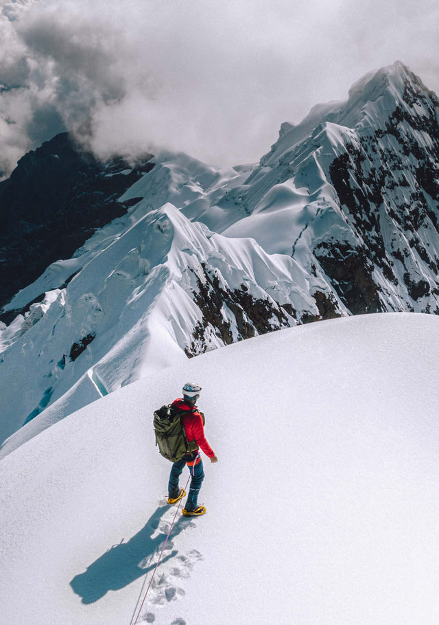Mountaineer in a red jacket climbing a snowy mountain ridge toward jagged peaks under a cloudy sky
