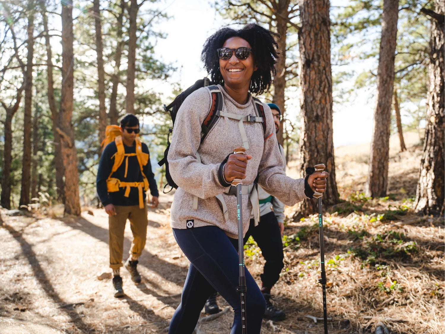 Woman hiking in relaxed-fit oatmeal alpaca-merino sweater, smiling and using trekking poles on a sunlit pine-forest trail