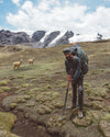 Hiker wearing an Ultralight Crew sweater hiking across an alpine meadow with backpack and trekking pole, cloudy mountains in the background