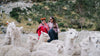 Two herders wearing red and blue jackets standing among a herd of white alpacas on a rocky Andean hillside