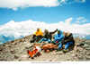 Two climbers resting at a high altitude basecamp with backpacks on a rocky ridge and snow covered peaks beyond