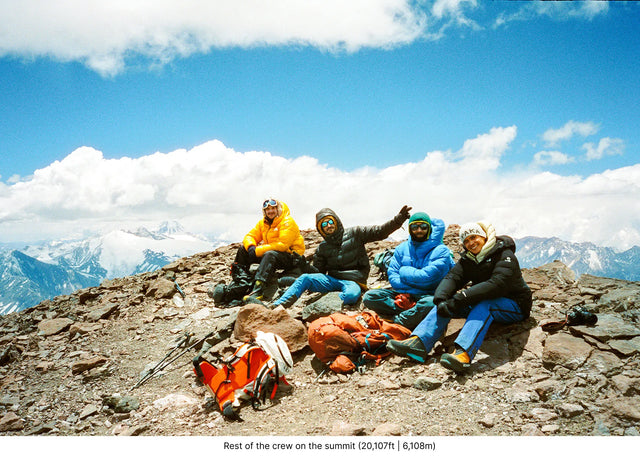 Two climbers resting at a high altitude basecamp with backpacks on a rocky ridge and snow covered peaks beyond