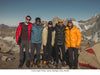 Five climbers standing on a rocky mountain slope near basecamp with snow-covered peaks and a clear blue sky