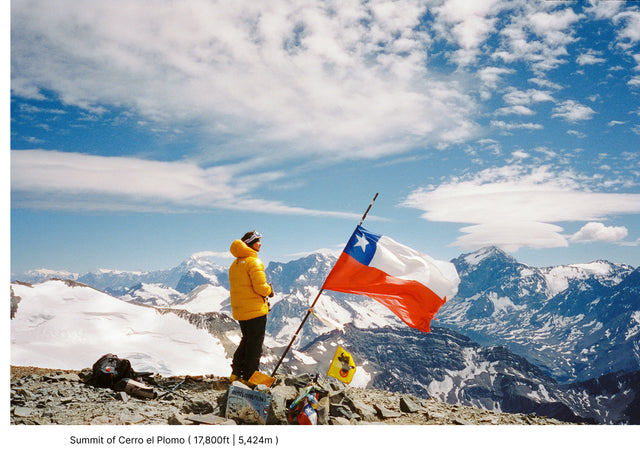 Two climbers on a snowy summit holding a Chilean flag with mountains and sky behind them
