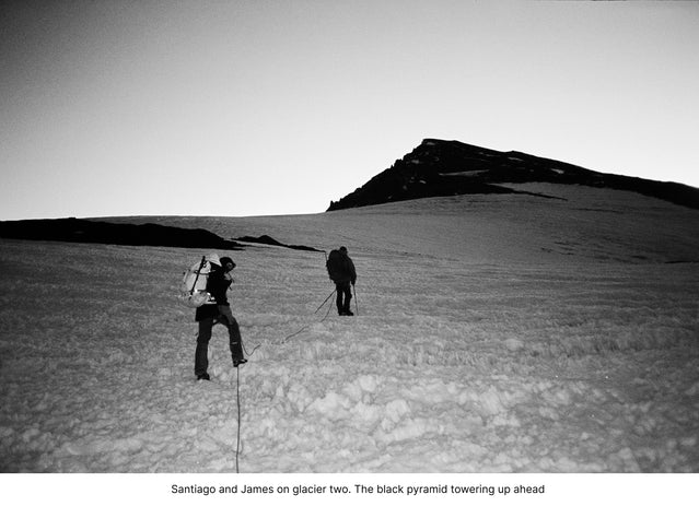 Two climbers walking toward a distant snow-covered peak across an alpine slope under a blue sky