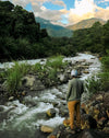 Hiker standing on a rocky riverbank looking toward forested mountains under a cloudy sky