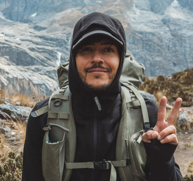Hiker wearing a black zip hoodie with a scuba-style hood and a cap, carrying a green backpack and flashing a peace sign on a mountain trail