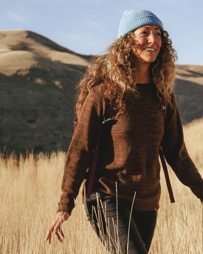 Woman walking on a sunlit grassy hillside wearing a brown cocoa alpaca hoodie in a relaxed fit, with a blue beanie and backpack