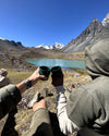 Two hikers clinking cups while seated beside a turquoise glacial lake with Ausangate mountains in the background