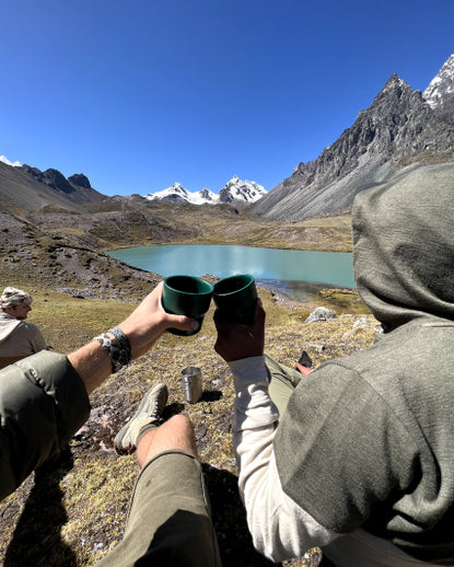Two hikers clinking cups while seated beside a turquoise glacial lake with Ausangate mountains in the background