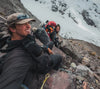 Three hikers resting on a rocky ledge beneath snow covered Ausangate peaks, wearing helmets and cold weather gear