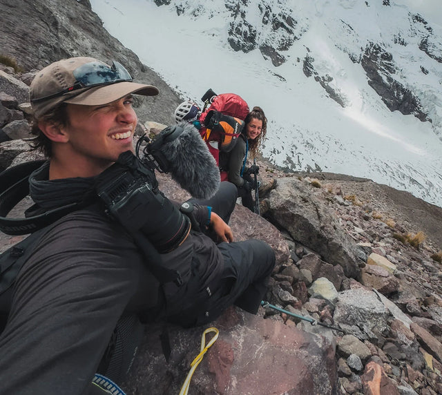 Three hikers resting on a rocky ledge beneath snow covered Ausangate peaks, wearing helmets and cold weather gear