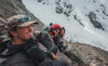 Two hikers taking a break on a rocky ridge, one holding a camera with a furry microphone, snowy mountain face and glacier in the background
