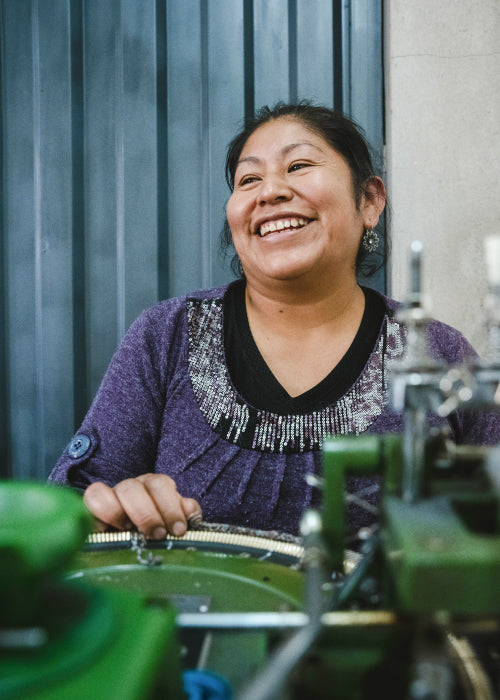 Woman wearing a purple Vida sweater smiling while operating a knitting machine in a workshop