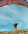 Person launching a red-orange paraglider above a grassy slope under a blue sky