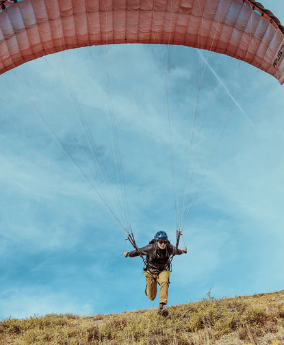Person launching a red-orange paraglider above a grassy slope under a blue sky