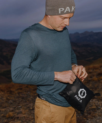 Person wearing teal Mayu ultralight alpaca jacket and PAKA beanie adjusting a camera outdoors at dusk