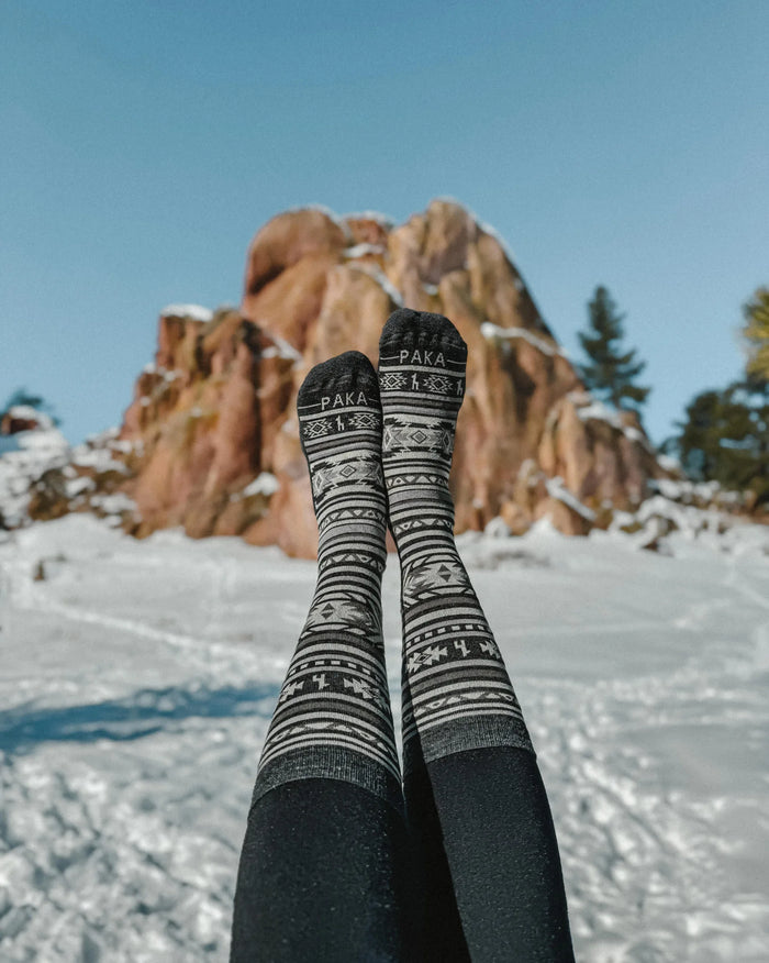 Legs wearing charcoal Inca crew socks with PAKA logo and Inca pattern, feet raised against snow and a rocky backdrop