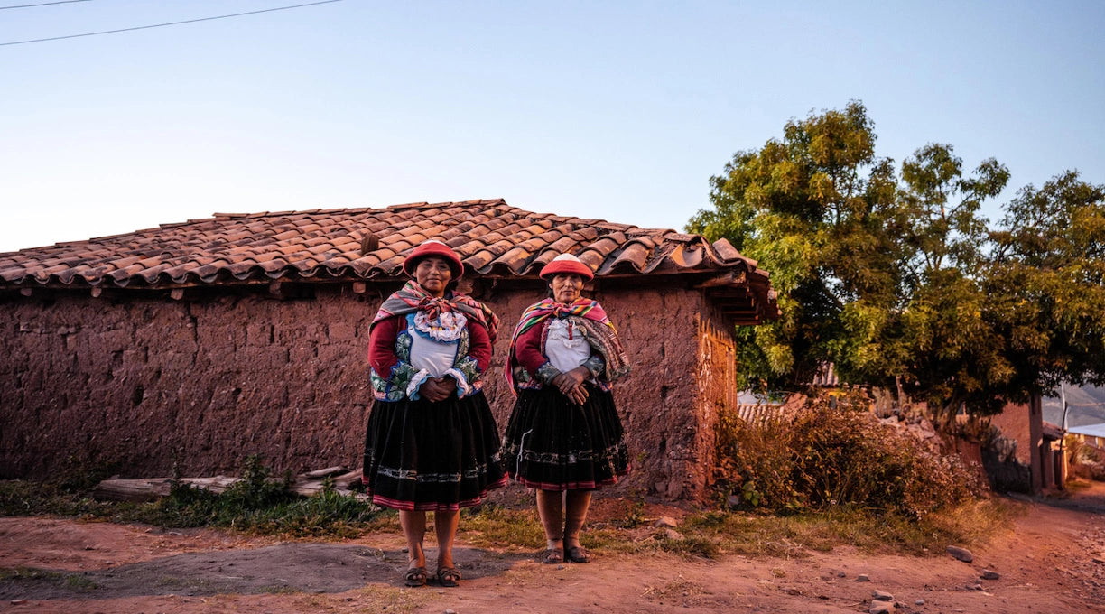 Two women in colorful traditional Andean clothing standing outside a low adobe house at sunset