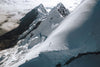 Climber in a red jacket ascending a steep snow ridge on Yanapaccha in the Cordillera Blanca, clouds below and jagged peaks behind
