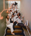 Group of teammates posing on an indoor stairway, smiling and waving with neon alpaca wall art in the background