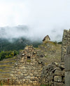 Stone ruins at Machu Picchu sit beneath misty Andean mountains, showing terraced agriculture and a small stone hut