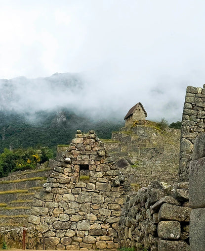 Stone ruins at Machu Picchu sit beneath misty Andean mountains, showing terraced agriculture and a small stone hut