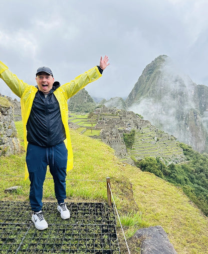 Person in yellow jacket raising arms on a misty mountain trail with stone terraces in the background